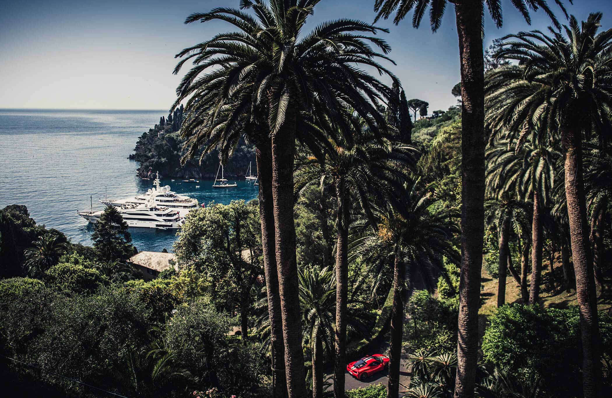 Red Ferrari driving along the South of France coastline with palm trees in the foreground and a super yacht in the background