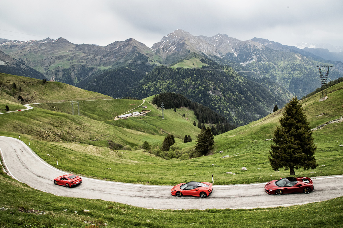 Three red Ferrari sports cars driving on a winding road through bright green alpine meadows and hills, with jagged grey mountains and a cloudy sky in the background.