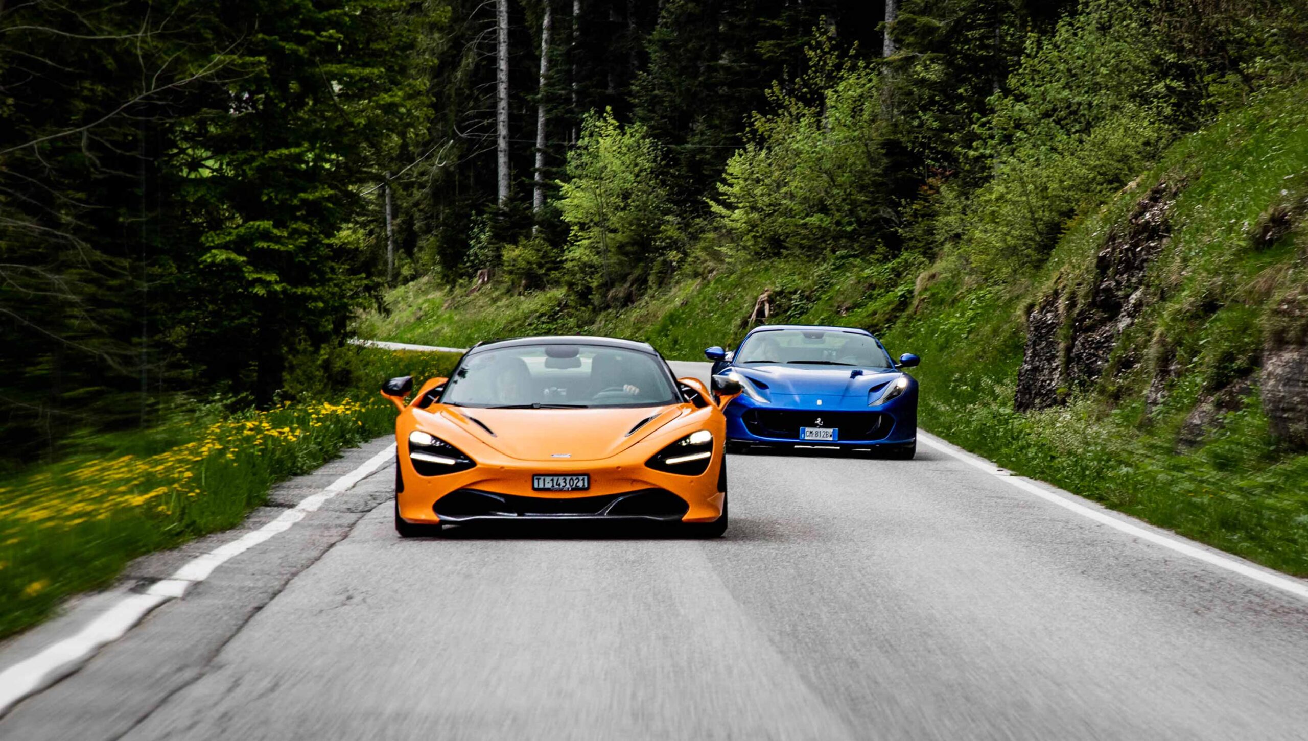 An orange McLaren sports car drives towards the camera on a two-lane road, closely followed by a blue Ferrari. The road is lined with lush green trees and grassy banks dotted with yellow wildflowers.