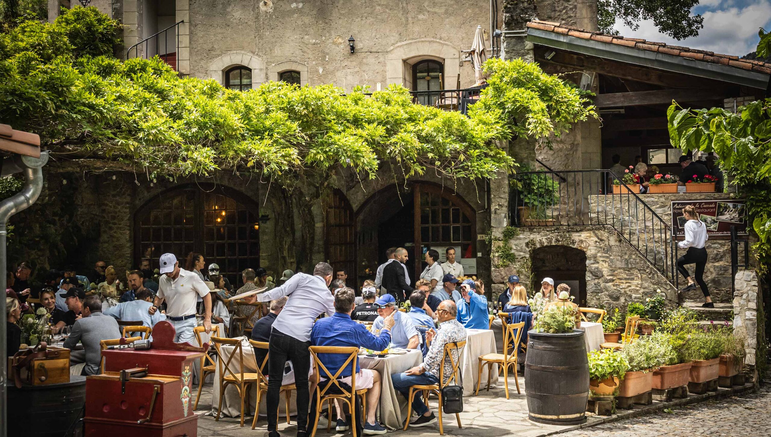 A busy outdoor restaurant patio is filled with diners seated at tables under the shade of a large, leafy green vine trellis. Waiters attend to guests in the courtyard of a rustic stone building.